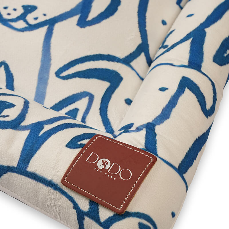 Close-up detail of the Paw Pop Bed showing the stitched DODO Pet Care logo label on a soft cream fabric with blue hand-drawn dog illustrations, highlighting premium stitching and plush texture.