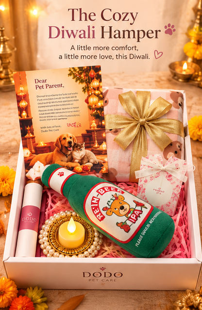 A Dodo Pet Care 'The Cozy Diwali Hamper' gift box filled with pet treats and toys, featuring a green and white 'Beer Bottle' plush toy, a pink gift box with a gold ribbon, a small white box with pink floral print, a lip balm tube, and a pet parent letter card, all arranged in pink shredded paper with a lit diya in the foreground.